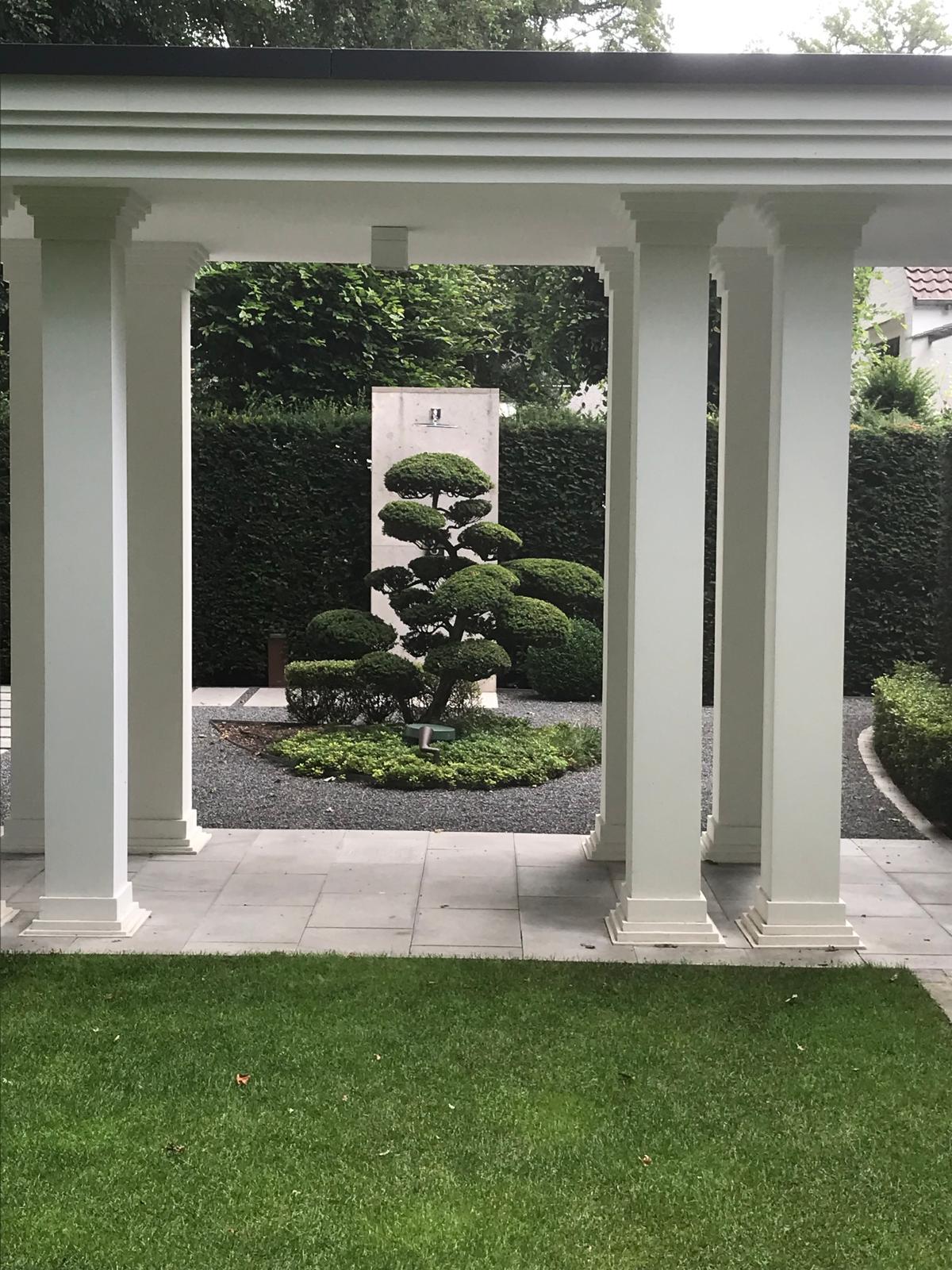 Blick durch eine weiße Pergola auf eine formgeschnittene Kiefer vor einer Wasserwand, gerahmt von hohen geschnittenen Hecken
